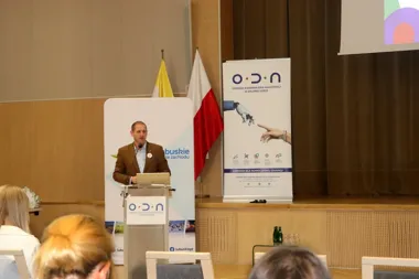 Mężczyzna w brązowej marynarce przemawia z pulpitu podczas konferencji w sali wykładowej, za nim stoją flagi i banery ODN oraz Lubuskie.